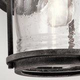 Close-up of the Ashland Bay Outdoor 1 Light Small Wall Lantern showcasing a colonial design. This lantern features a clear, seeded glass enclosure with charming bubbles that beautifully complements the weathered zinc metal frame encasing the lamp. A light bulb is visible inside.