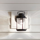 A charming Colonial-style outdoor wall lantern from Ashland Bay, featuring a clear glass shade and a black metal frame with a weathered zinc finish, is mounted on a white horizontal paneling exterior wall. The light casts a warm glow and soft shadows, perfectly embodying rustic design aesthetics.