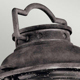 Close-up of the Ashland Bay Outdoor 1 Light Medium Pedestal Lantern top, featuring a textured weathered zinc finish. The lantern includes a curved handle and visible rivets, with a portion of the glass and metal frame beneath. The background is a soft light gray.