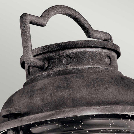 Close-up of the Ashland Bay Outdoor 1 Light Medium Pedestal Lantern top, featuring a textured weathered zinc finish. The lantern includes a curved handle and visible rivets, with a portion of the glass and metal frame beneath. The background is a soft light gray.