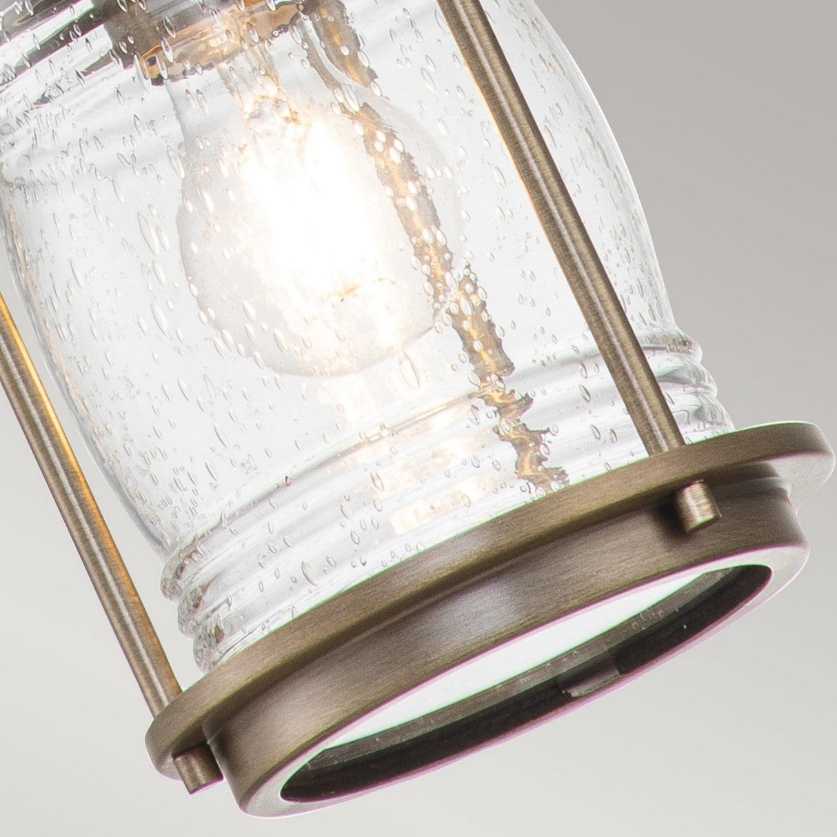Close-up of the Ashland Bay Outdoor 1 Light Small Chain Lantern, reminiscent of a Colonial Style Lantern. It features a clear, textured glass shade showcasing an illuminated bulb inside and metallic accents in burnished bronze. The background is a plain, light gray color.