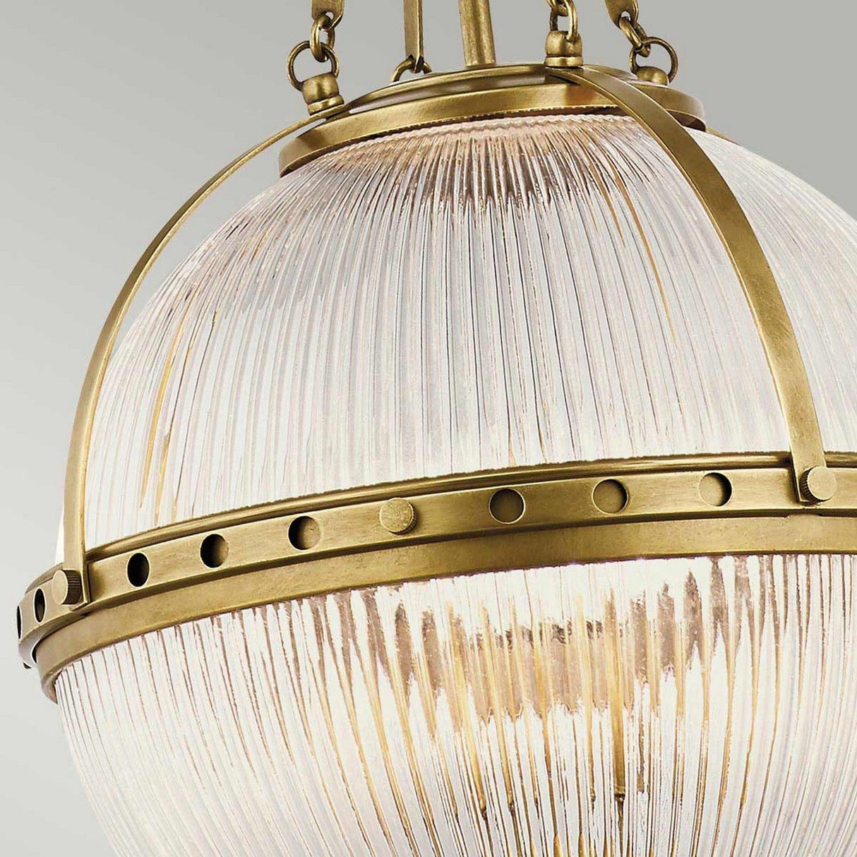 Get a close-up view of the Aster 3 Light Pendant - Brass, showcasing its vintage-style charm with ribbed glass domes and an antique brass finish. Decorative rivets enhance the metal band surrounding the glass, elegantly suspended by a chain.
