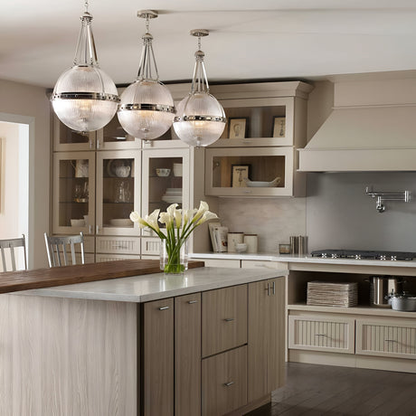The modern kitchen boasts a spacious island with a light wood finish and white countertop, illuminated by three Aster 3 Light Pendant fixtures in Polished Nickel, each featuring ribbed glass domes. Cabinets with glass doors showcase an array of dishes, while a vase of white calla lilies adorns the island. Stainless steel appliances in Polished Nickel add to the sleek aesthetic.