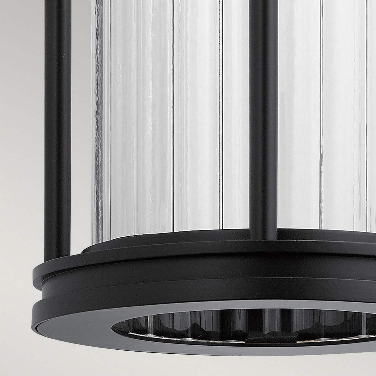 Close-up of the Barras Outdoor 1 Light Medium Wall Lantern, featuring a cylindrical ribbed glass body encased in a matte black metal frame. The clear glass enhances the lantern's sleek and contemporary design.