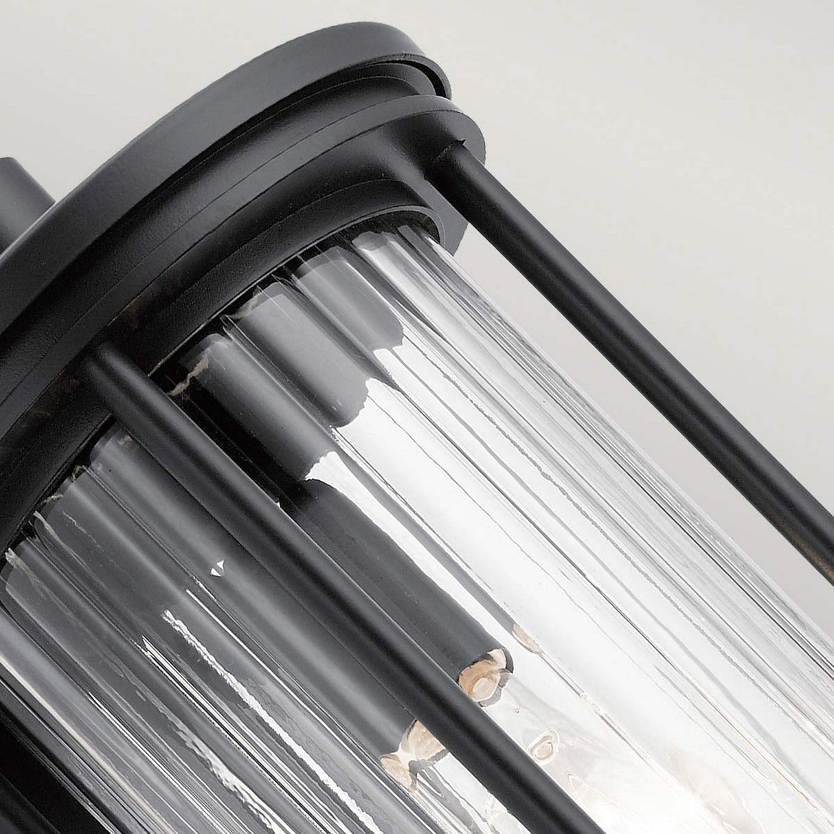 A detailed view of the Barras Outdoor 1 Light Medium Wall Lantern in black, featuring a cylindrical design with clear glass and vertical metal bars that expose the internal components. The ribbed glass adds texture to the fixture, set against a plain background.