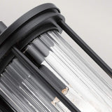 A detailed view of the Barras Outdoor 1 Light Medium Wall Lantern in black, featuring a cylindrical design with clear glass and vertical metal bars that expose the internal components. The ribbed glass adds texture to the fixture, set against a plain background.