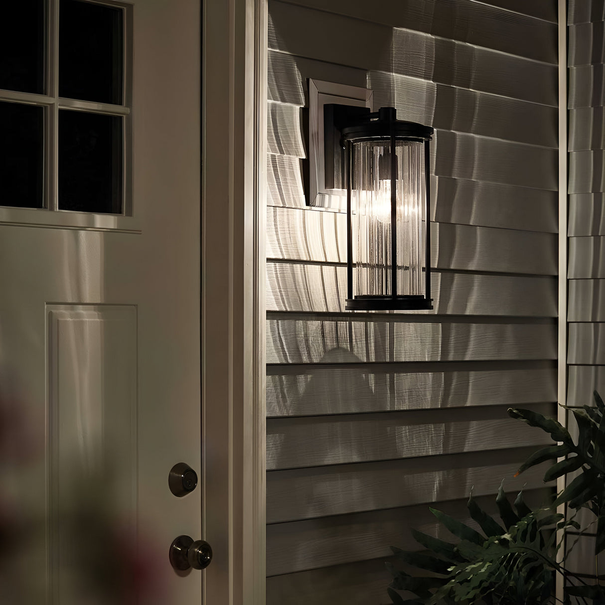 The Barras Outdoor 1 Light Medium Wall Lantern in black, featuring ribbed glass, casts intricate shadows on the house siding at night. A closed white door with a small window is visible to the left, while leaves in the lower right corner enhance the charm of this outdoor lighting fixture.