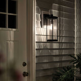The Barras Outdoor 1 Light Medium Wall Lantern in black, featuring ribbed glass, casts intricate shadows on the house siding at night. A closed white door with a small window is visible to the left, while leaves in the lower right corner enhance the charm of this outdoor lighting fixture.