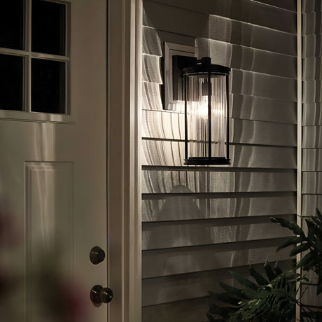 The Barras Outdoor 1 Light Medium Wall Lantern in black, featuring ribbed glass, casts intricate shadows on the house siding at night. A closed white door with a small window is visible to the left, while leaves in the lower right corner enhance the charm of this outdoor lighting fixture.