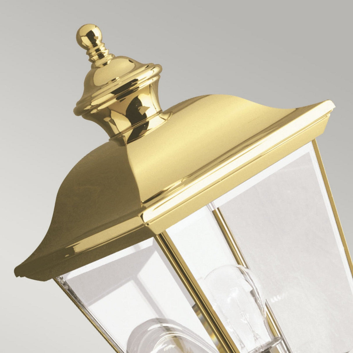Close-up of the Bay Shore Outdoor 1 Light Medium Outdoor Wall Light in polished brass, showcasing its decorative top. The lantern features clear beveled glass panels and two visible light bulbs, set against a plain gray background.