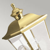 Close-up of the Bay Shore Outdoor 1 Light Medium Outdoor Wall Light in polished brass, showcasing its decorative top. The lantern features clear beveled glass panels and two visible light bulbs, set against a plain gray background.