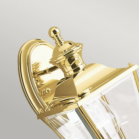 Close-up of the Bay Shore Outdoor 1 Light Small Outdoor Wall Light in polished brass, showcasing its classic design. The lantern features bevelled glass panels and an ornate top with a finial, while its curved mount gleams against a gray background.
