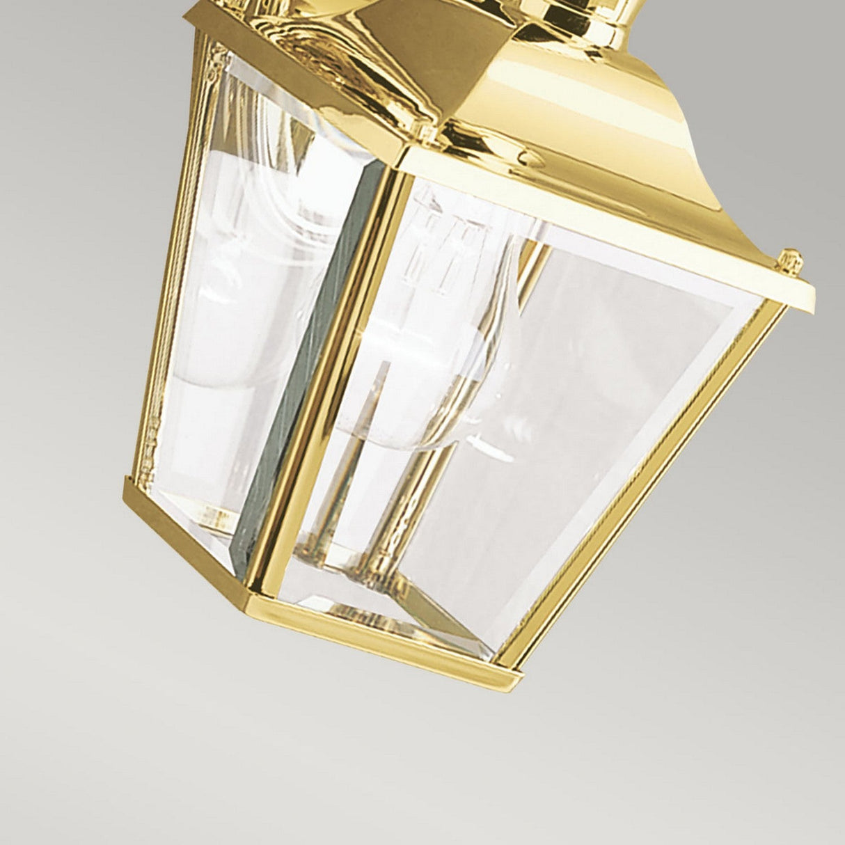 A close-up of the Bay Shore Outdoor 1 Light Small Outdoor Wall Light in polished brass showcases its bevelled glass panels and visible light bulb against a plain gray background.