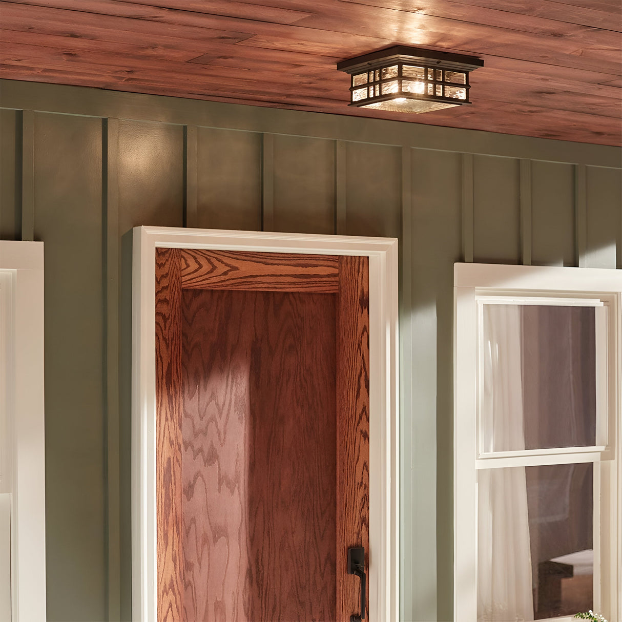 The exterior of the home highlights its craftsmen architecture, featuring a wooden door flanked by two windows with white trim. Above the door, an Olde Bronze-finished light fixture from the Beacon Outdoor 2 Light Flush collection enhances the sage green wall with a warm glow.
