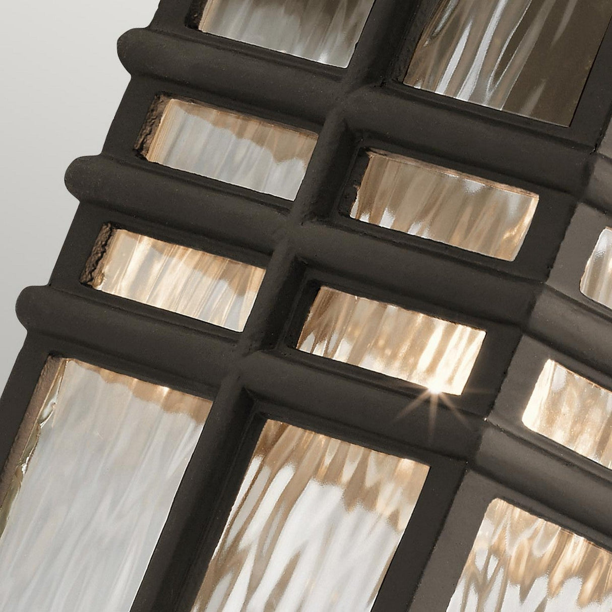 Close-up of the Beacon Square Outdoor 1 Light Medium Wall Lantern in bronze, showcasing its geometric, grid-like metal frame with textured glass panels. The warm light shines through the glass, creating a soft glow and highlighting the intricate design. The lantern is durable and weather-resistant with its IP44 rating.