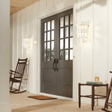 A cozy porch showcases craftsman-inspired design with a pair of dark double doors adorned with glass panels. Two wooden rocking chairs sit on either side, one accompanied by a drink on a nearby table. The entrance is warmly illuminated by the Beacon Square Outdoor 1 Light Small Wall Lantern in white, and completed with a UV-resistant doormat for an inviting touch.