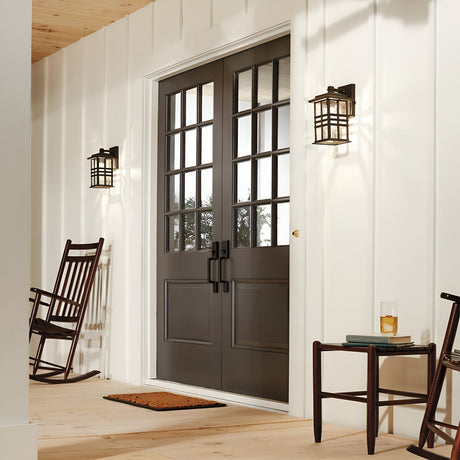A cozy porch showcases dark double doors with glass panes, complemented by the Beacon Square Outdoor 1 Light Small Wall Lantern in bronze, which enhances the craftsman-inspired design. On one side, a wooden rocking chair invites relaxation, while on the other side, a small table holds a glass of beverage and a book. A doormat lies invitingly at the entrance.