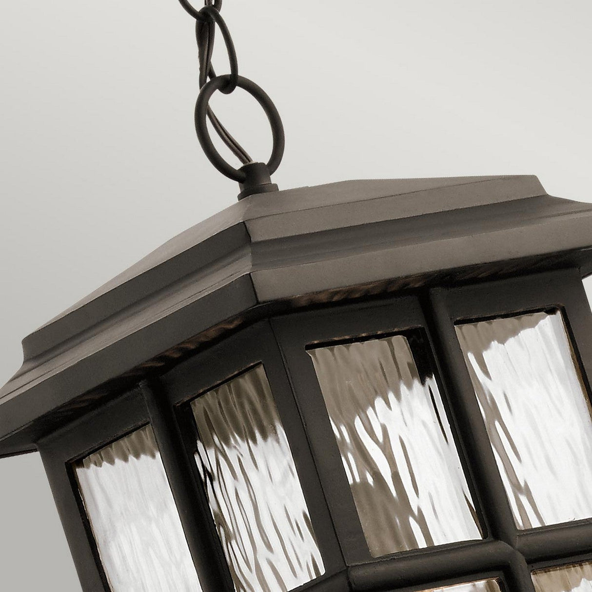 A close-up of the Beacon Square Outdoor 1 Light Chain Lantern - Bronze shows its textured glass panels gleaming atop a neutral backdrop. The lantern's dark metal frame and bronze finish create an elegant display as it hangs gracefully from a simple chain.