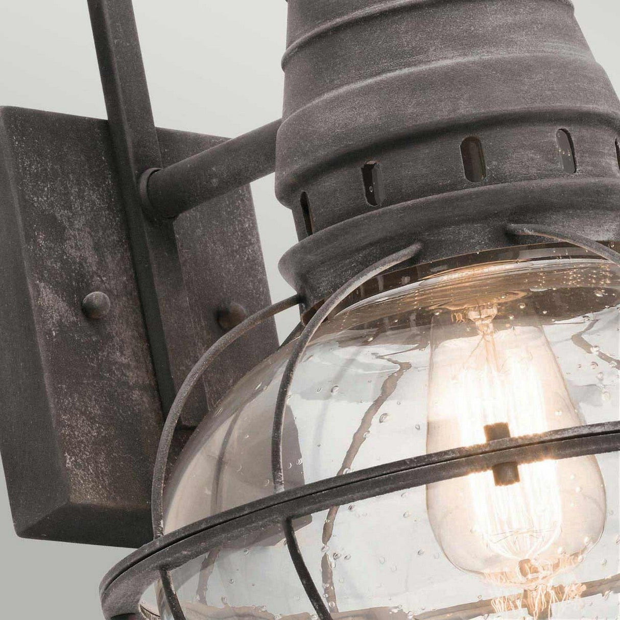 A close-up of the Bridgepoint Outdoor 1 Light Large Wall Lantern highlights its clear glass shade and visible Edison bulb. This outdoor lighting fixture features a weathered zinc finish with raindrops on the glass, providing a rustic and aged look.
