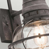 A close-up of the Bridgepoint Outdoor 1 Light Large Wall Lantern highlights its clear glass shade and visible Edison bulb. This outdoor lighting fixture features a weathered zinc finish with raindrops on the glass, providing a rustic and aged look.