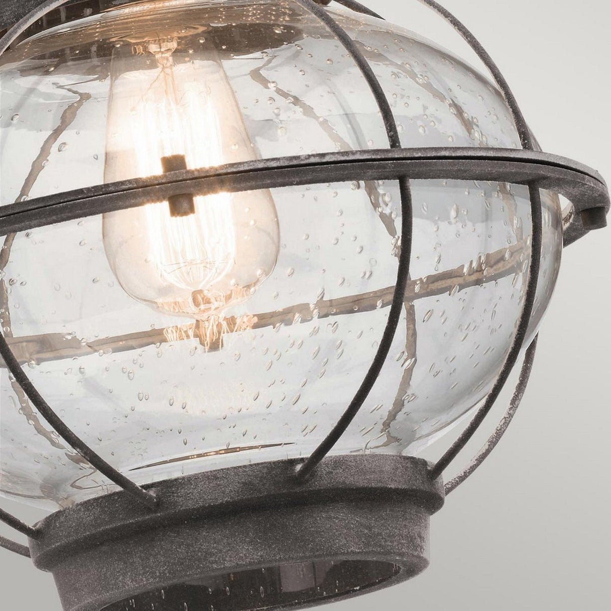 Close-up of the Bridgepoint Outdoor 1 Light Large Wall Lantern - Weathered Zinc featuring a vintage-style light bulb inside a black metal cage. The speckled, rain-like glass and weathered zinc finish enhance its charm, as the warm glow highlights the lantern's rustic design against a light gray background.