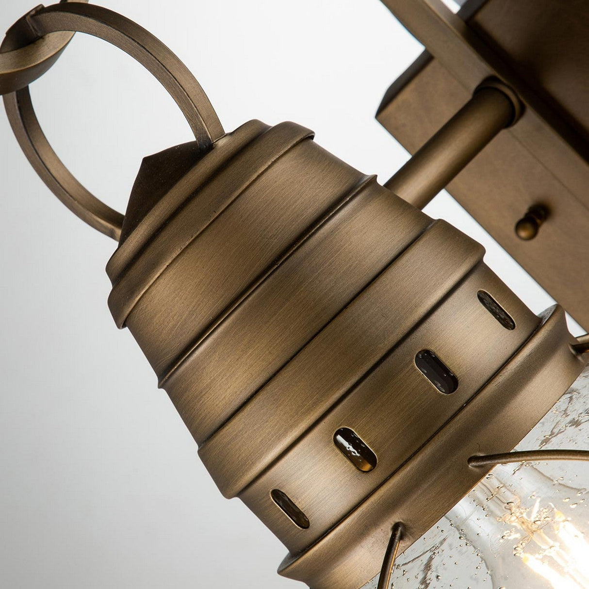 A close-up of the Bridgepoint Outdoor 1 Light Medium Wall Lantern in natural brass, featuring a classic, tiered design. The metal fixture encases a clear glass shade, highlighting a visible light bulb. Its nautical style brings charming elegance to any exterior space.