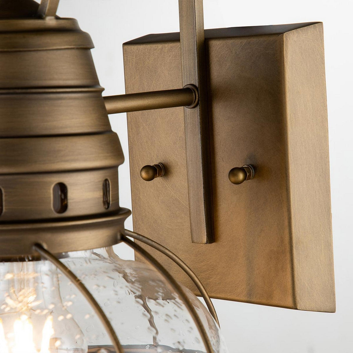 A close-up of the Bridgepoint Outdoor 1 Light Medium Wall Lantern in natural brass showcases its vintage-style charm. The fixture boasts a clear glass shade encasing the bulb, offering a hint of nautical style. Its square base is embellished with two small decorative knobs that enhance its timeless appeal.