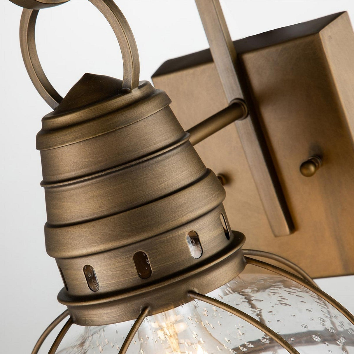 A detailed view of the Bridgepoint Outdoor 1 Light Medium Wall Lantern in natural brass showcases its vintage charm. The fixture includes a clear, textured glass shade encompassed by a cage-like frame and is mounted on a square brass plate. Its design exudes an industrial, nautical style, making it ideal as an outdoor wall lantern or indoor accent piece.