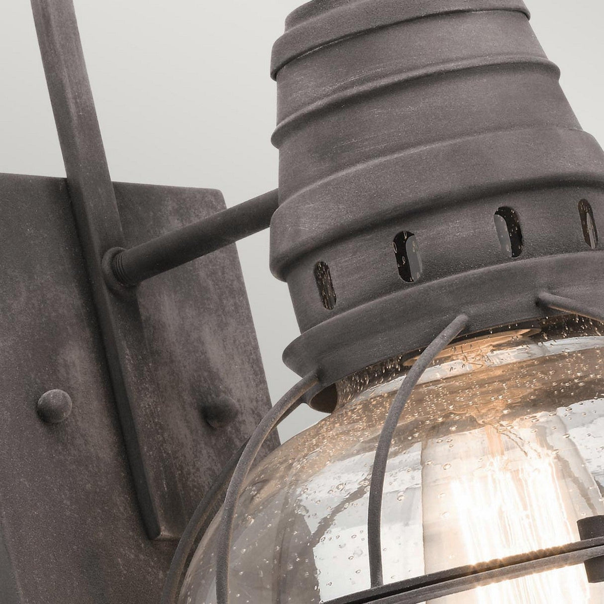 Close-up of the Bridgepoint Outdoor 1 Light Medium Wall Lantern with a weathered zinc finish. It showcases a rustic metal frame and seeded glass cover, revealing a visible bulb that emits a warm light. The finish contributes to its vintage aesthetic, enhancing its charm with an antique feel.