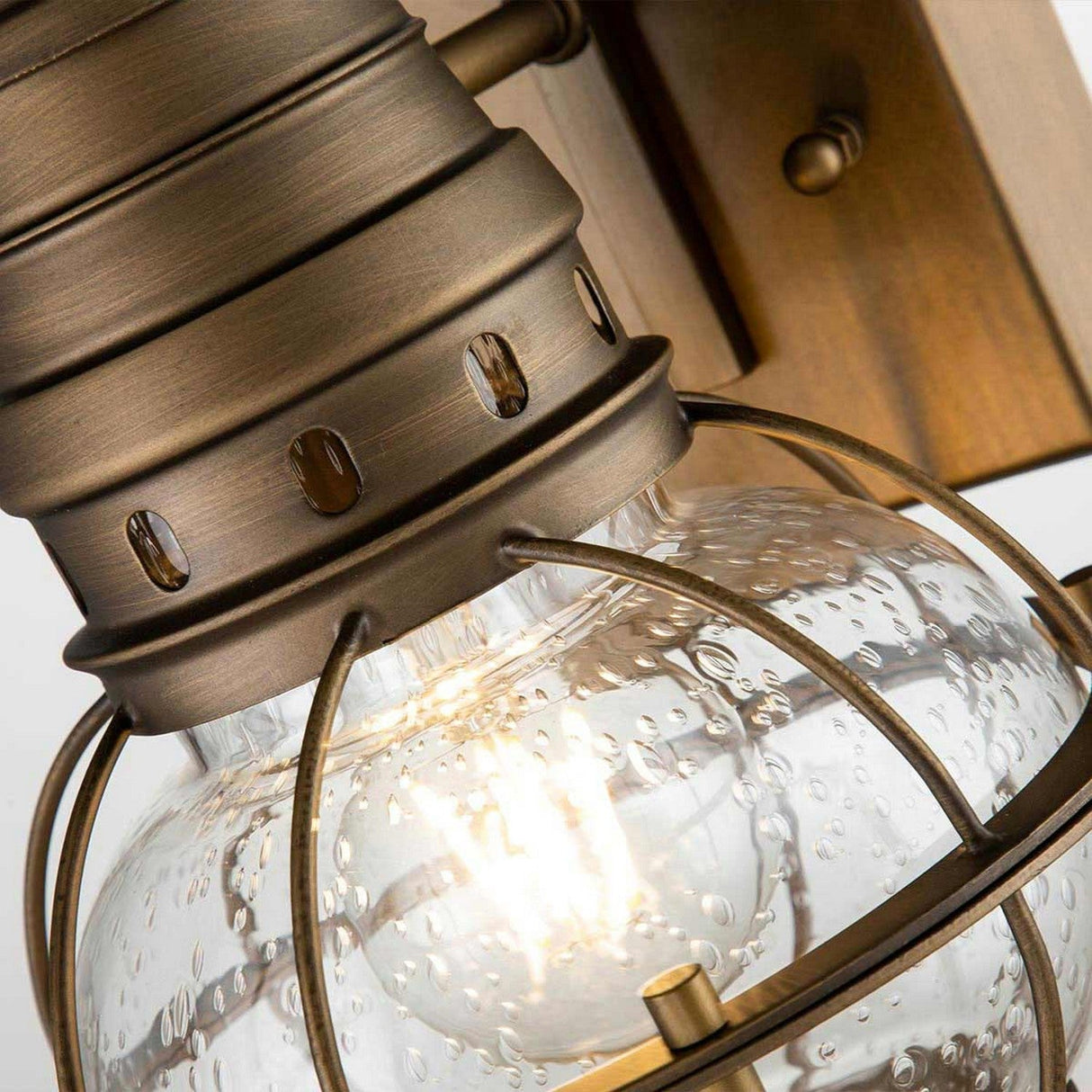 Get a close-up view of the Bridgepoint Outdoor 1 Light Small Wall Lantern in natural brass, showcasing its bronze metal frame and clear glass globe with a water droplet texture. Ideal for nautical outdoor lighting, this fixture houses a warm glowing bulb inside that highlights its intricate details beautifully.