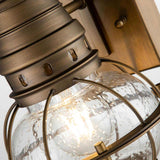 Get a close-up view of the Bridgepoint Outdoor 1 Light Small Wall Lantern in natural brass, showcasing its bronze metal frame and clear glass globe with a water droplet texture. Ideal for nautical outdoor lighting, this fixture houses a warm glowing bulb inside that highlights its intricate details beautifully.