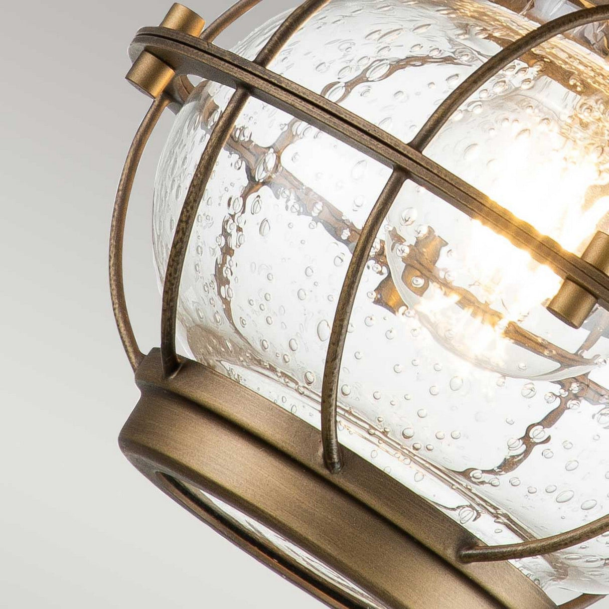 Close-up of the Bridgepoint Outdoor 1 Light Small Wall Lantern in Natural Brass, featuring a clear, textured glass globe encased in a bronze metal cage. A warm glow emanates from within, accentuating the bubble details in the glass.