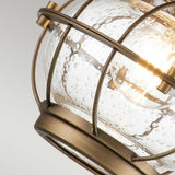 Close-up of the Bridgepoint Outdoor 1 Light Small Wall Lantern in Natural Brass, featuring a clear, textured glass globe encased in a bronze metal cage. A warm glow emanates from within, accentuating the bubble details in the glass.