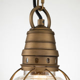 An up-close view of the Bridgepoint Outdoor 1 Light Small Chain Lantern showcases its vintage design with a natural brass finish. It features a cylindrical top adorned with circular cutouts and is complemented by a clear seeded glass shade, supported by metal brackets. The fixture is equipped with a black cord for easy hanging.