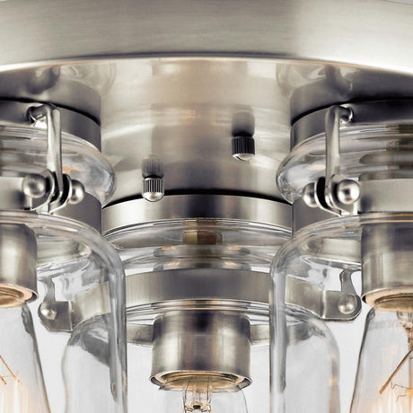 Close-up of the Brinley 3 Light Flush in brushed nickel, showcasing its modern design with clear glass shades encircling each bulb. The fixture exudes a sleek, industrial style with visible screws and clamps, accentuating its polished finish and expert craftsmanship.