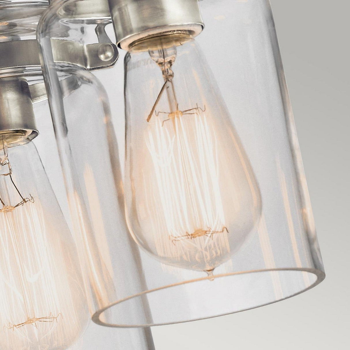 A close-up of the Brinley 3 Light Flush shows vintage-style Edison bulbs enclosed in clear glass shades, casting a warm, soft glow. This fixture features a brushed nickel attachment at the top, effortlessly combining modern design with classic appeal for vintage-inspired lighting.