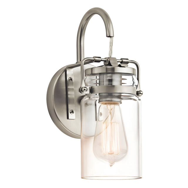 The Brinley 1 Light Wall Light - Brushed Nickel is a vintage-style wall sconce featuring an exposed filament bulb enclosed in a clear glass jar. Its design, which includes a curved arm connecting the fixture to a round base plate, perfectly captures vintage appeal.