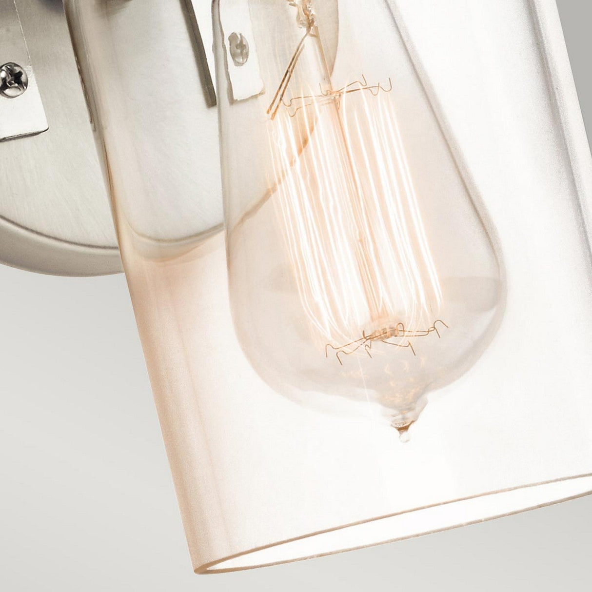 A close-up of the Brinley 1 Light Wall Light showcases a vintage-style bulb with an exposed filament encased in clear glass, attached to a brushed nickel fixture. The warm glow beautifully contrasts against a neutral background, highlighting the bulb's intricate design and vintage charm.