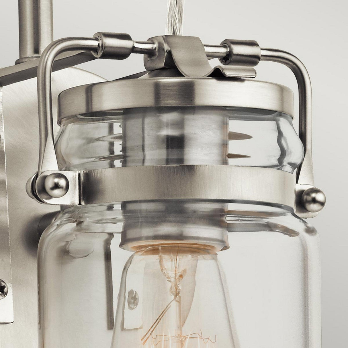 Close-up of the Brinley 1 Light Wall Light - Brushed Nickel, showcasing its vintage charm with a glass and metal design. The visible light bulb is encased within the glass jar, held securely by brushed nickel brackets against a softly lit plain background.