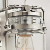 Close-up of the Brinley 1 Light Wall Light - Brushed Nickel, showcasing its vintage charm with a glass and metal design. The visible light bulb is encased within the glass jar, held securely by brushed nickel brackets against a softly lit plain background.