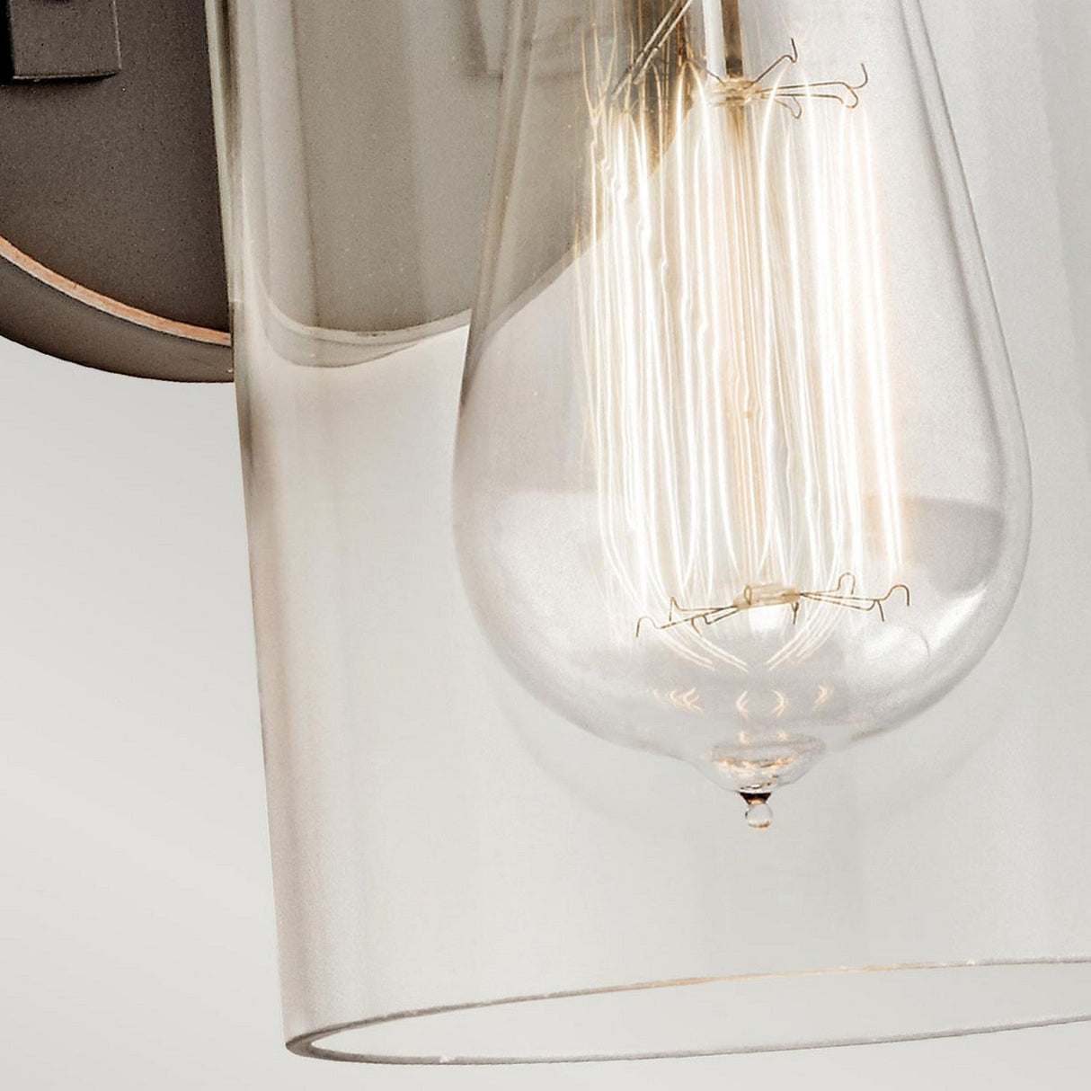 A close-up of the Brinley 1 Light Wall Light in bronze showcases its vintage design with an industrial-style bulb featuring visible filaments. Encased in a cylindrical glass shade, the illuminated bulb casts a warm glow against a neutral background, perfectly complementing the fixture's classic charm.