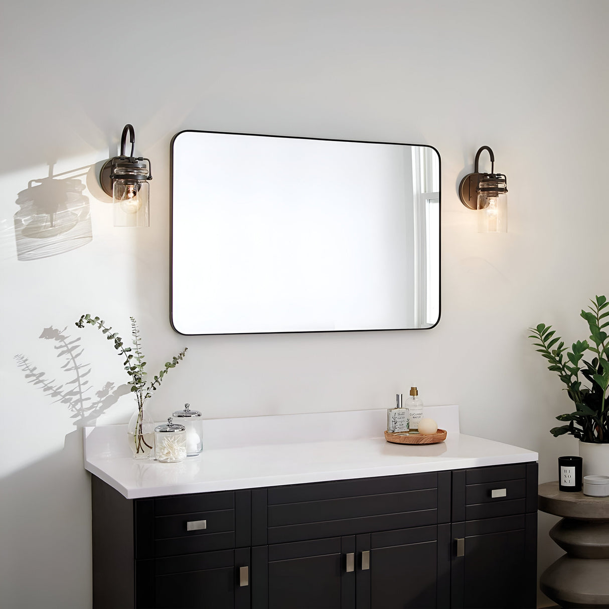 A modern bathroom showcases a black vanity with a white countertop. Above it, a large rectangular mirror is accompanied by Brinley 1 Light Wall Lights in bronze, featuring clear glass shades. Potted plants and bathroom essentials decorate the space.