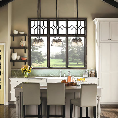 A modern kitchen with a vintage-inspired touch features a large window, the Brinley 6 Light Pendant in bronze finish, and a central island. White cabinets, shelves displaying dishes, and a glass pitcher filled with drinks complete the look, while a vase of yellow tulips adorns the countertop.