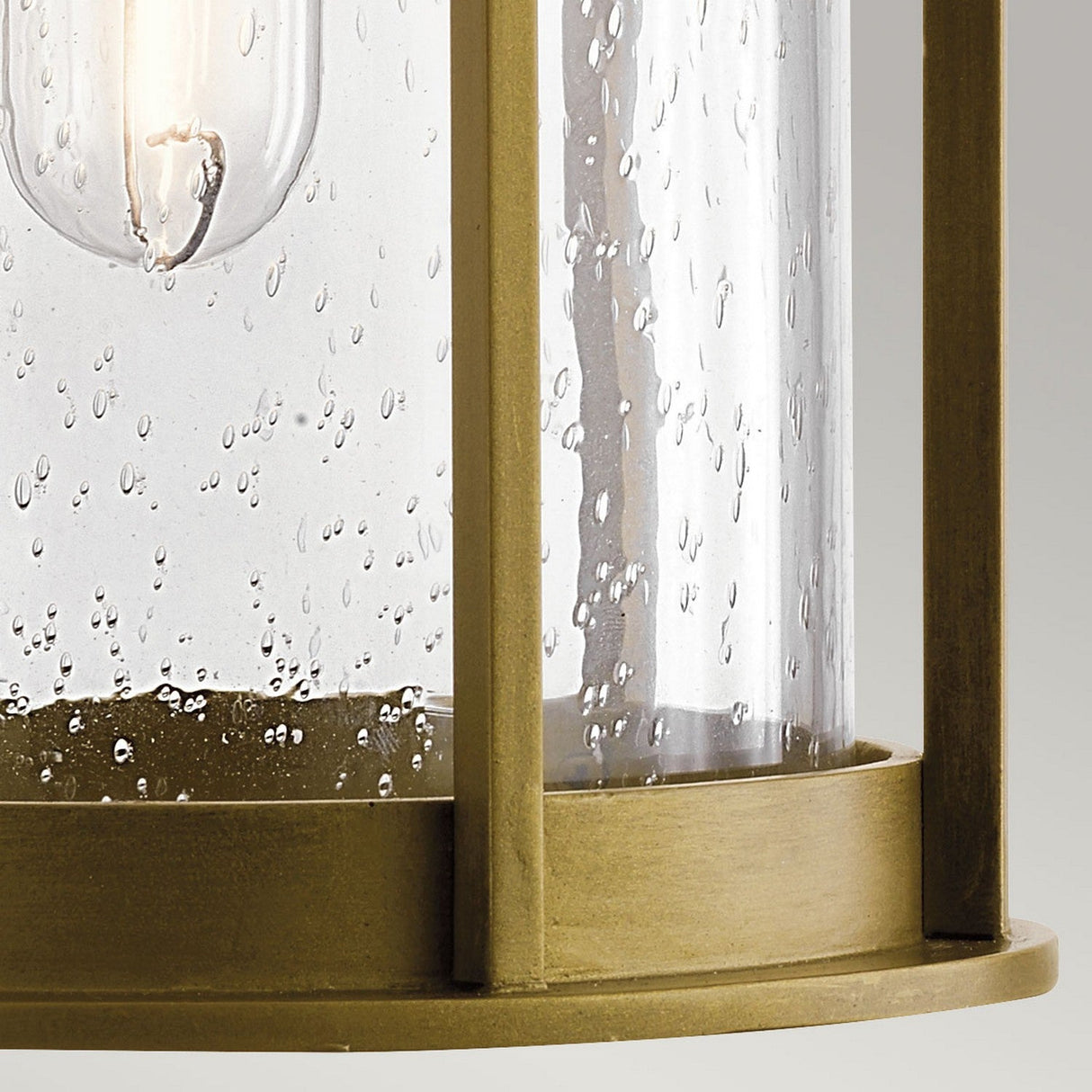 The Camillo Outdoor 1 Light Medium Wall Lantern - Natural Brass features a close-up of a lantern-style light fixture, complete with a natural brass frame and clear seeded glass panels. The decorative bubbles in the glass offer glimpses of the glowing bulb inside, enhancing its warm ambiance.