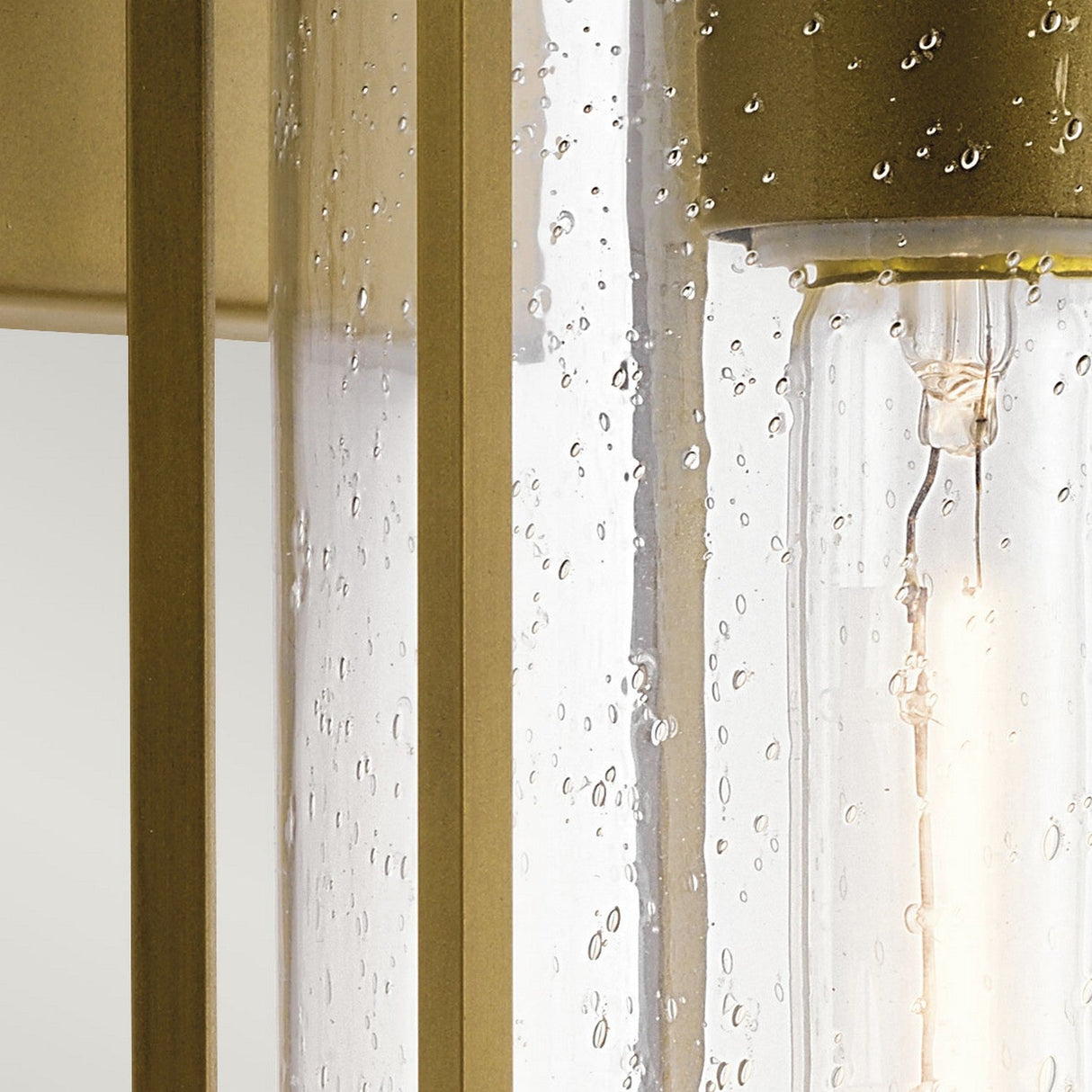 Close-up of the Camillo Outdoor 1 Light Medium Wall Lantern, showcasing its raindrop-like texture similar to clear seeded glass. Inside, a visible light bulb emits a soft glow. Framed in natural brass metal, this lantern exudes elegance and modernity, making it an ideal piece for outdoor lighting against a neutral background.