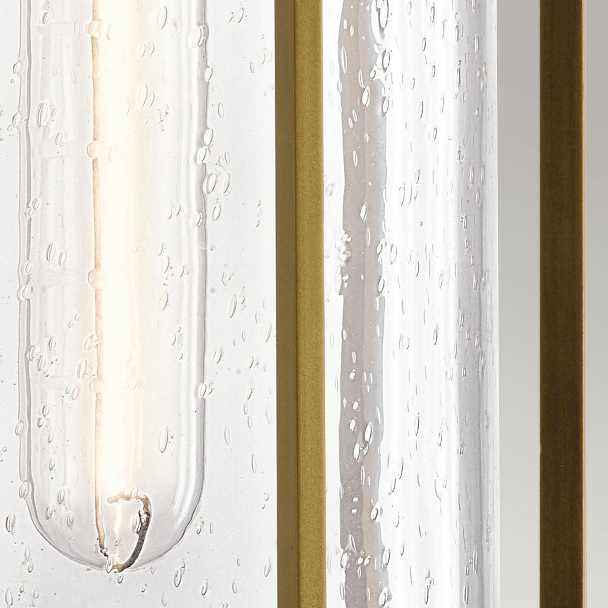 A close-up of the Camillo Outdoor 1 Light Medium Wall Lantern in Natural Brass showcasing clear seeded glass adorned with water droplets and bordered by vertical golden lines. The natural brass accents combined with the gentle light filtering through its slightly transparent surface offer a warm and elegant ambiance.