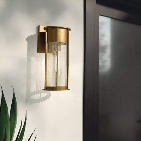 The Camillo Outdoor 1 Light Medium Wall Lantern, crafted in natural brass, tastefully enhances a white wall. Its sleek panels of clear seeded glass capture the sunlight beautifully, casting graceful shadows. A hint of lush green foliage peeks through at the bottom left corner, infusing a touch of nature into this sophisticated scene.
