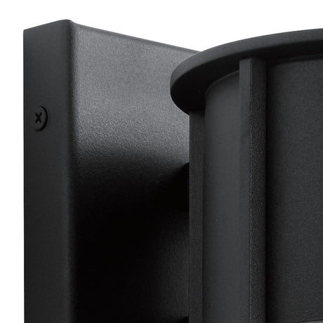 Close-up of the Camillo Outdoor 1 Light Medium Wall Lantern in a sleek black finish, featuring a cylindrical design. Mounted on a square metal base, it showcases clear seeded glass with a visible screw on the side. The fixture possesses a sophisticated matte surface that enhances any contemporary exterior.