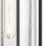 A close-up view of the Camillo Outdoor 1 Light Medium Wall Lantern - Black reveals a clear seeded glass tube adorned with water droplets. Inside, a glowing filament emits a warm glow, framed by an elegant black finish, beautifully showcasing the texture and transparency of the glass.