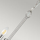 A close-up of the Capitol Hill 12 Light Chandelier in brushed nickel showcases its modern, industrial-style design. The fixture features a sleek rod and chain with candle-shaped bulbs that have clear filaments encased in metal holders, all set against a neutral background.